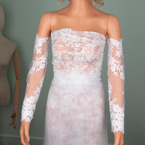 Puede incluir: Vestido de novia de encaje blanco exhibido en un maniqu&iacute;. El vestido presenta un corpi&ntilde;o sin tirantes y mangas largas y transparentes con detalles de encaje floral. La falda es un dise&ntilde;o largo y fluido. El estilo general es elegante y rom&aacute;ntico.