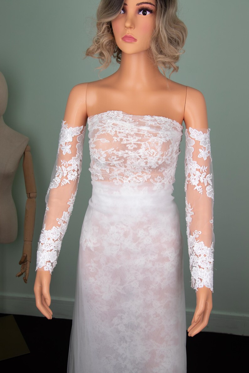 Puede incluir: Vestido de novia de encaje blanco exhibido en un maniqu&iacute;. El vestido presenta un corpi&ntilde;o sin tirantes y mangas largas y transparentes con detalles de encaje floral. La falda es un dise&ntilde;o largo y fluido. El estilo general es elegante y rom&aacute;ntico.