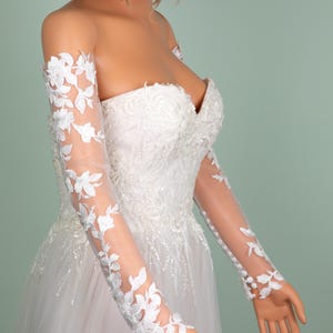 May include: A strapless white wedding dress with a sweetheart neckline and a full tulle skirt. The dress features intricate floral lace detailing and sheer long sleeves adorned with matching floral appliques. The dress is displayed on a mannequin.