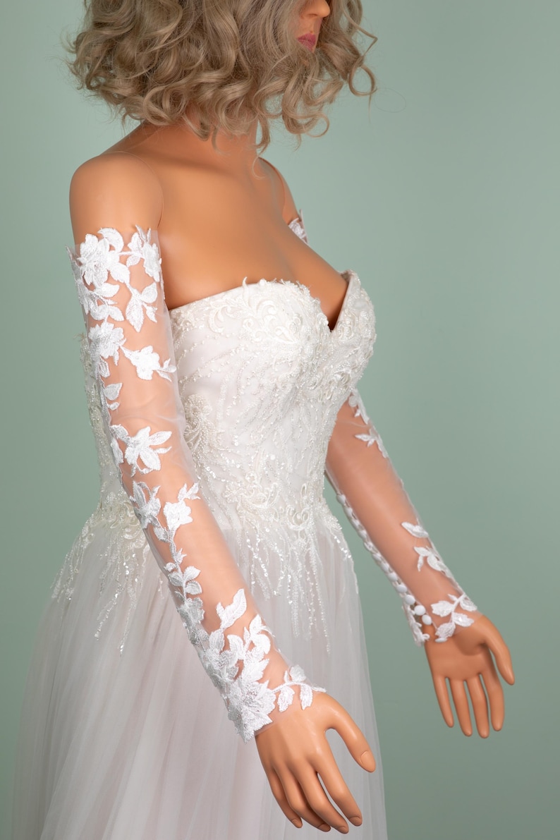 May include: A strapless white wedding dress with a sweetheart neckline and a full tulle skirt. The dress features intricate floral lace detailing and sheer long sleeves adorned with matching floral appliques. The dress is displayed on a mannequin.