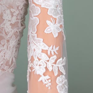 Puede incluir: Primer plano de una manga de vestido de novia de encaje blanco. La manga est&aacute; adornada con bordados florales y lentejuelas, creando un dise&ntilde;o delicado y elegante. La tela transparente revela la piel debajo.