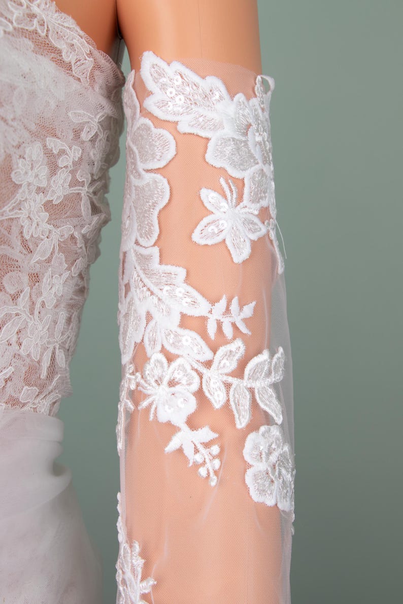 Puede incluir: Primer plano de una manga de vestido de novia de encaje blanco. La manga est&aacute; adornada con bordados florales y lentejuelas, creando un dise&ntilde;o delicado y elegante. La tela transparente revela la piel debajo.