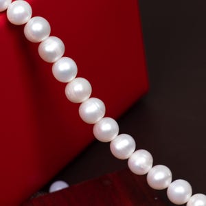 Puede incluir: Un collar de perlas blancas y brillantes sobre un fondo rojo. Las perlas son redondas y de tamaño uniforme, ensartadas para formar un collar. El fondo es rojo intenso, con una zona marrón oscuro en la esquina.