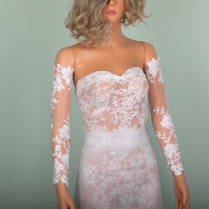 Peut inclure: Robe de mari&eacute;e en dentelle blanche sans bretelles avec de longues manches transparentes. La robe pr&eacute;sente des d&eacute;tails de dentelle florale complexes et un d&eacute;collet&eacute; en c&oelig;ur. Les manches sont orn&eacute;es d'applications florales assorties.