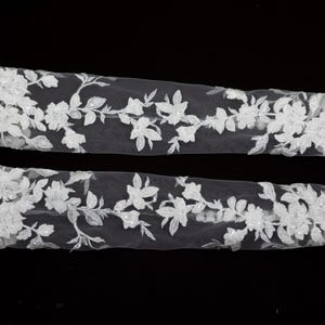 May include: Two strips of white floral lace with intricate detailing. The lace features a delicate pattern of flowers and leaves, perfect for embellishing bridal wear or other craft projects.