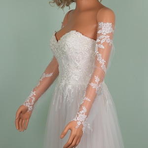 May include: A white strapless wedding dress with intricate floral embroidery on the bodice and a flowing tulle skirt. Sheer, long sleeves with delicate white floral lace detailing are attached. The dress is displayed on a mannequin.