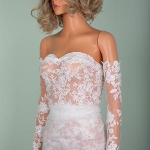 Peut inclure: Robe de mari&eacute;e blanche sans bretelles avec des d&eacute;tails de dentelle florale. La robe pr&eacute;sente des manches longues et transparentes avec des accents floraux et une superposition de dentelle assortie sur le corsage et la jupe. Une ceinture blanche cintre la taille.
