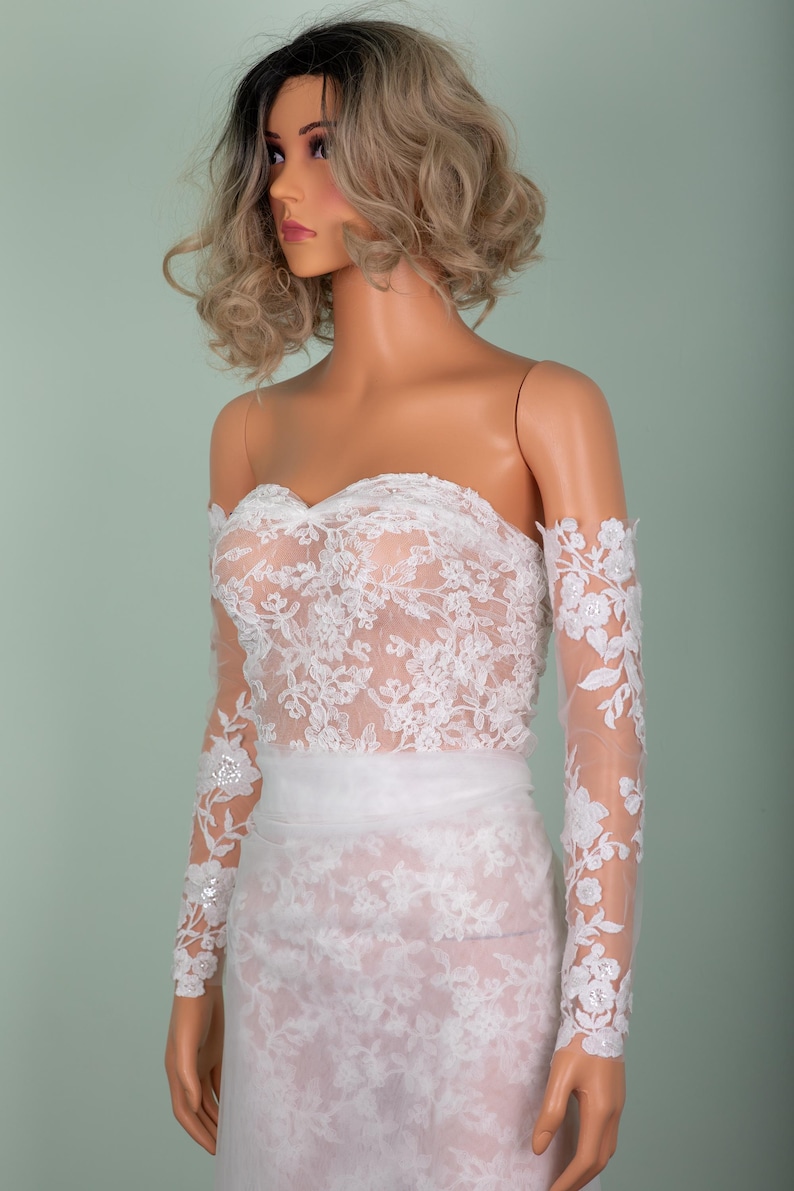 Peut inclure: Robe de mari&eacute;e blanche sans bretelles avec des d&eacute;tails de dentelle florale. La robe pr&eacute;sente des manches longues et transparentes avec des accents floraux et une superposition de dentelle assortie sur le corsage et la jupe. Une ceinture blanche cintre la taille.