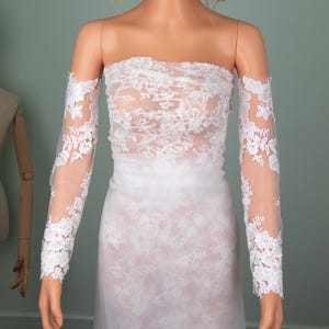 Puede incluir: Un vestido de novia blanco sin tirantes con delicados detalles de encaje. El vestido presenta un corpi&ntilde;o ajustado y una falda fluida. Tambi&eacute;n se incluyen mangas largas y transparentes de encaje, que a&ntilde;aden un toque elegante al vestido de novia. El vestido se exhibe en un maniqu&iacute;.