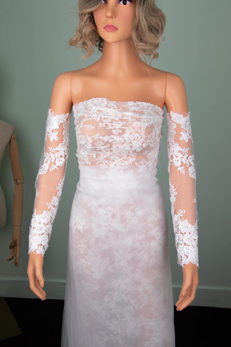 Puede incluir: Un vestido de novia blanco sin tirantes con delicados detalles de encaje. El vestido presenta un corpi&ntilde;o ajustado y una falda fluida. Tambi&eacute;n se incluyen mangas largas y transparentes de encaje, que a&ntilde;aden un toque elegante al vestido de novia. El vestido se exhibe en un maniqu&iacute;.