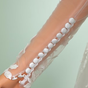 May include: Close-up of a sheer white sleeve with a row of white buttons. The sleeve is adorned with delicate lace detailing and floral patterns. The background is a soft, muted green.