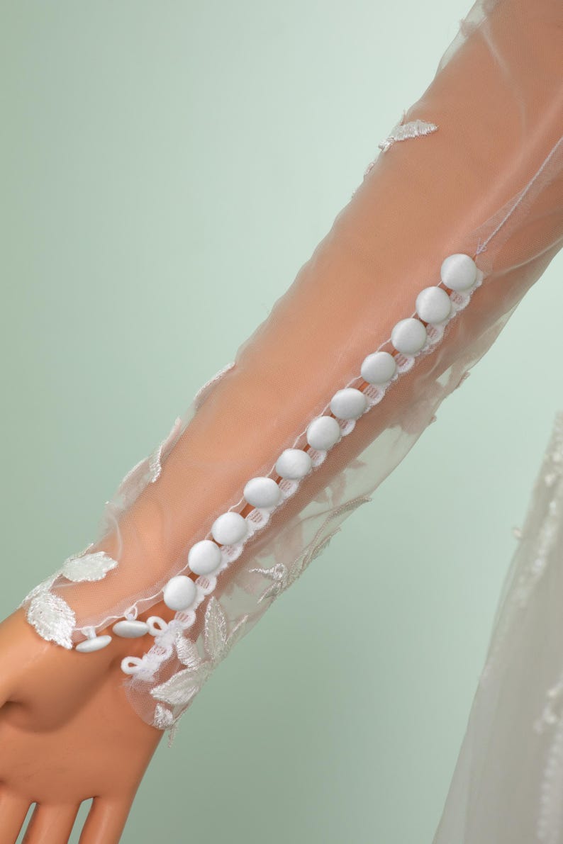 May include: Close-up of a sheer white sleeve with a row of white buttons. The sleeve is adorned with delicate lace detailing and floral patterns. The background is a soft, muted green.