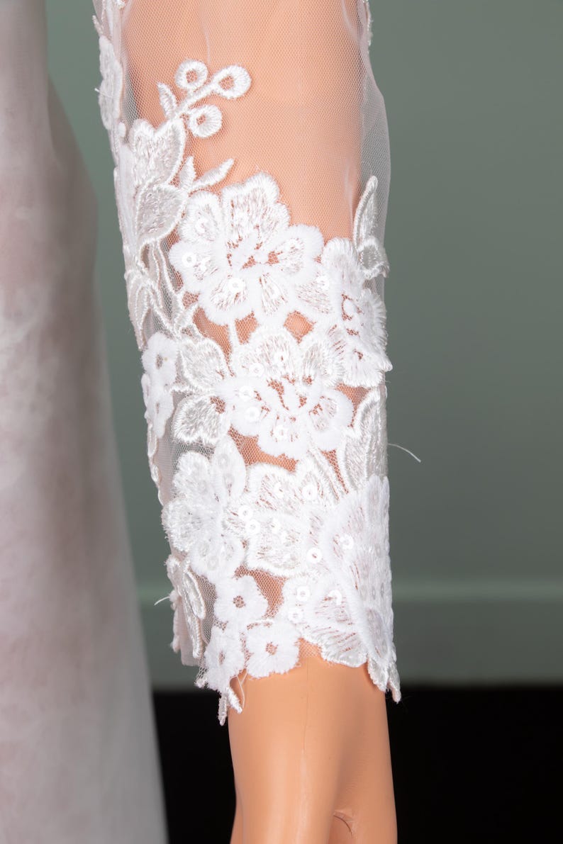 Puede incluir: Primer plano de una manga de encaje blanco. La manga presenta un intrincado bordado floral con un respaldo de malla transparente. El delicado dise&ntilde;o de encaje es detallado y elegante, lo que sugiere ropa de novia o formal.