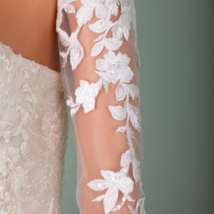 May include: Close-up of a white bridal gown with floral lace detailing. The sheer sleeve features delicate white floral embroidery with sequin accents. The dress is displayed on a mannequin against a soft green background.