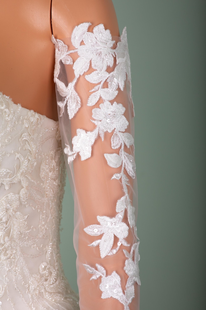 May include: Close-up of a white bridal gown with floral lace detailing. The sheer sleeve features delicate white floral embroidery with sequin accents. The dress is displayed on a mannequin against a soft green background.