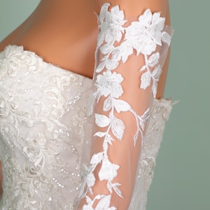 May include: A white wedding dress with intricate beadwork and floral embroidery. The sheer sleeve is adorned with delicate white floral lace, creating an elegant and romantic look. The dress is a strapless design, perfect for a special occasion.
