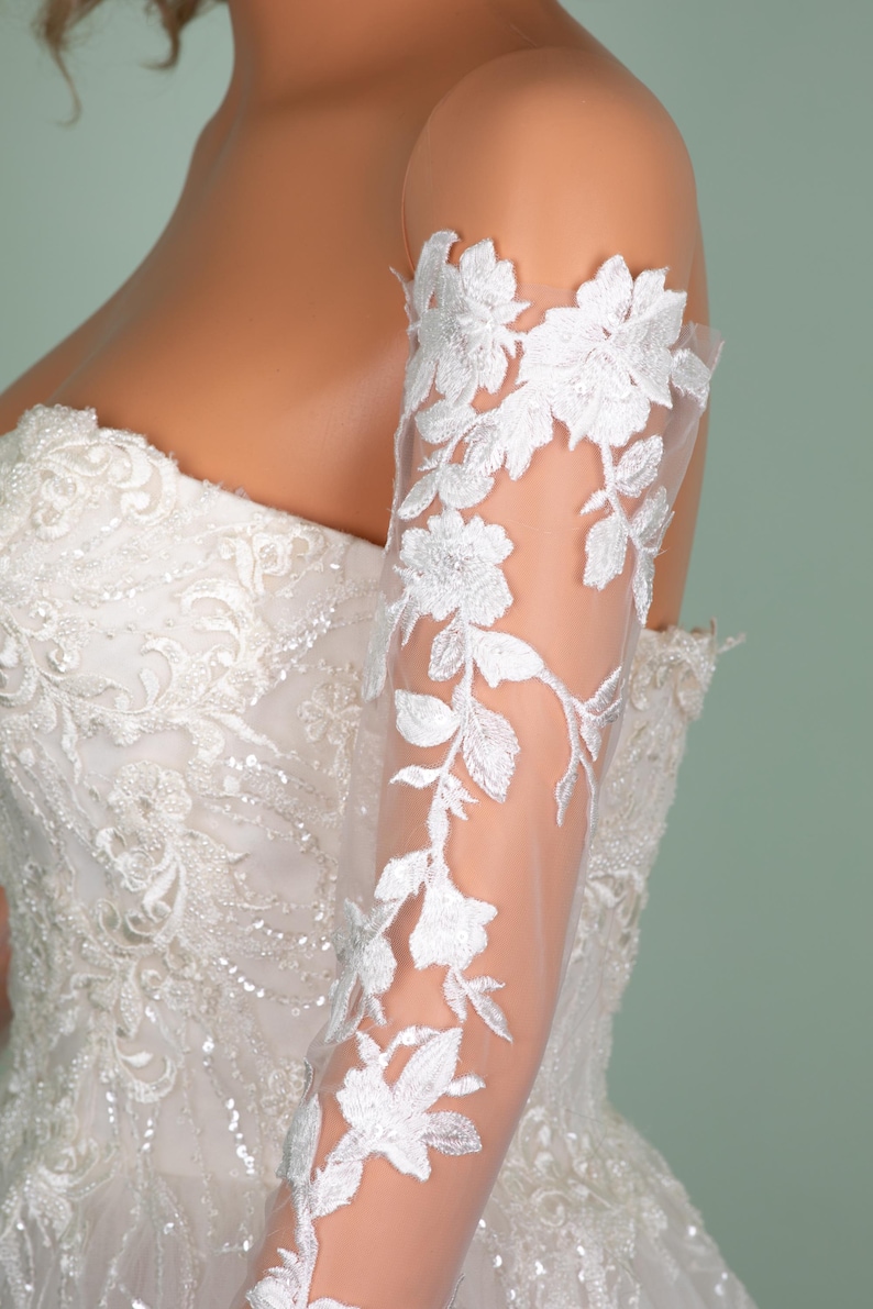 May include: A white wedding dress with intricate beadwork and floral embroidery. The sheer sleeve is adorned with delicate white floral lace, creating an elegant and romantic look. The dress is a strapless design, perfect for a special occasion.
