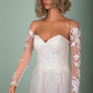 May include: A strapless white wedding dress with floral lace detailing and sheer long sleeves adorned with floral appliques. The dress features a sweetheart neckline and a flowing tulle skirt, displayed on a mannequin.
