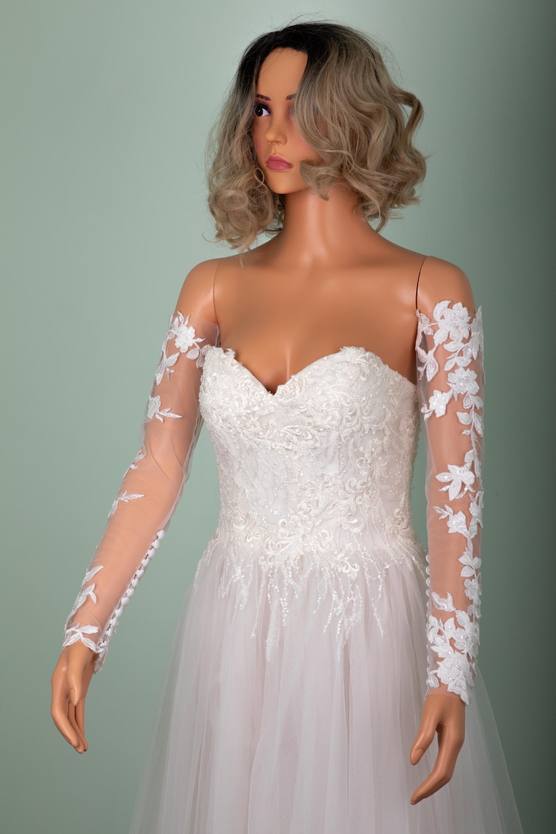 May include: A strapless white wedding dress with floral lace detailing and sheer long sleeves adorned with floral appliques. The dress features a sweetheart neckline and a flowing tulle skirt, displayed on a mannequin.