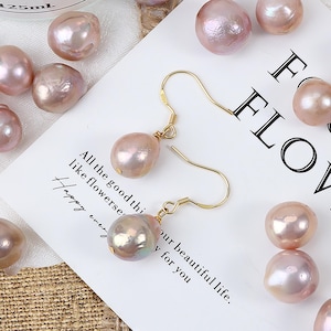 Könnte beinhalten: Ein Paar goldene Ohrringe mit rosafarbenen Barockperlen-Tropfen. Die Perlen sind irisierend und haben eine natürliche, unregelmäßige Form. Die Ohrringe werden auf einer weißen Oberfläche mit anderen losen Perlen präsentiert.