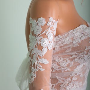 Peut inclure: Robe de mari&eacute;e en dentelle blanche avec broderie florale. La robe pr&eacute;sente une manche transparente avec des appliqu&eacute;s de dentelle florale d&eacute;taill&eacute;s. Le corsage et la manche sont orn&eacute;s de dentelle blanche d&eacute;licate, cr&eacute;ant un look &eacute;l&eacute;gant et romantique.