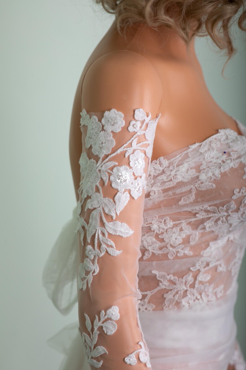 Peut inclure: Robe de mari&eacute;e en dentelle blanche avec broderie florale. La robe pr&eacute;sente une manche transparente avec des appliqu&eacute;s de dentelle florale d&eacute;taill&eacute;s. Le corsage et la manche sont orn&eacute;s de dentelle blanche d&eacute;licate, cr&eacute;ant un look &eacute;l&eacute;gant et romantique.
