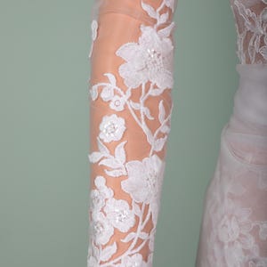Peut inclure: Gros plan sur une manche en dentelle florale blanche. La dentelle d&eacute;licate pr&eacute;sente des motifs d&eacute;taill&eacute;s de fleurs et de feuilles avec de subtils accents de paillettes. La manche est transparente, r&eacute;v&eacute;lant la peau en dessous, et fait partie d'une robe de mari&eacute;e.