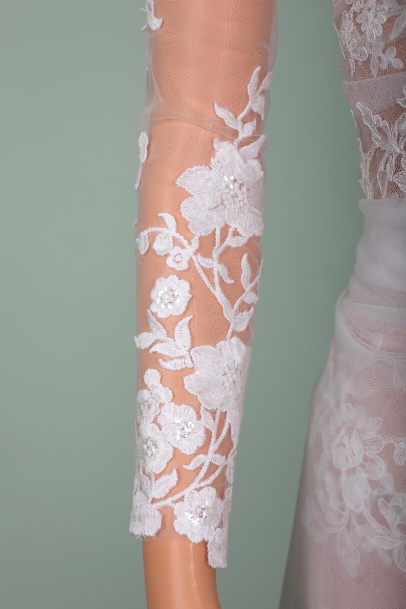 Peut inclure: Gros plan sur une manche en dentelle florale blanche. La dentelle d&eacute;licate pr&eacute;sente des motifs d&eacute;taill&eacute;s de fleurs et de feuilles avec de subtils accents de paillettes. La manche est transparente, r&eacute;v&eacute;lant la peau en dessous, et fait partie d'une robe de mari&eacute;e.