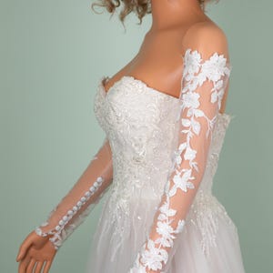May include: A white bridal gown with a strapless bodice and a full skirt. The sheer sleeves are adorned with delicate floral lace. The sleeves are buttoned at the wrist. The dress is embellished with intricate beadwork and embroidery.