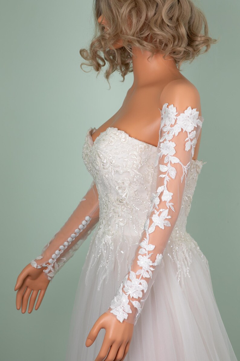 May include: A white bridal gown with a strapless bodice and a full skirt. The sheer sleeves are adorned with delicate floral lace. The sleeves are buttoned at the wrist. The dress is embellished with intricate beadwork and embroidery.