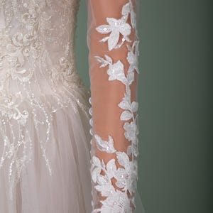May include: A white wedding dress with intricate floral lace detailing. The long sleeves are sheer, adorned with delicate white floral appliques and sequins. The bodice features detailed embroidery and beadwork, and the skirt is made of layers of tulle.