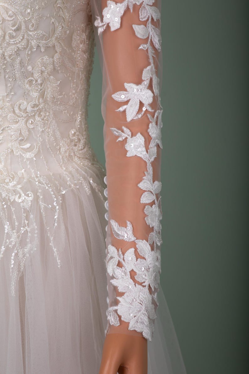 May include: A white wedding dress with intricate floral lace detailing. The long sleeves are sheer, adorned with delicate white floral appliques and sequins. The bodice features detailed embroidery and beadwork, and the skirt is made of layers of tulle.