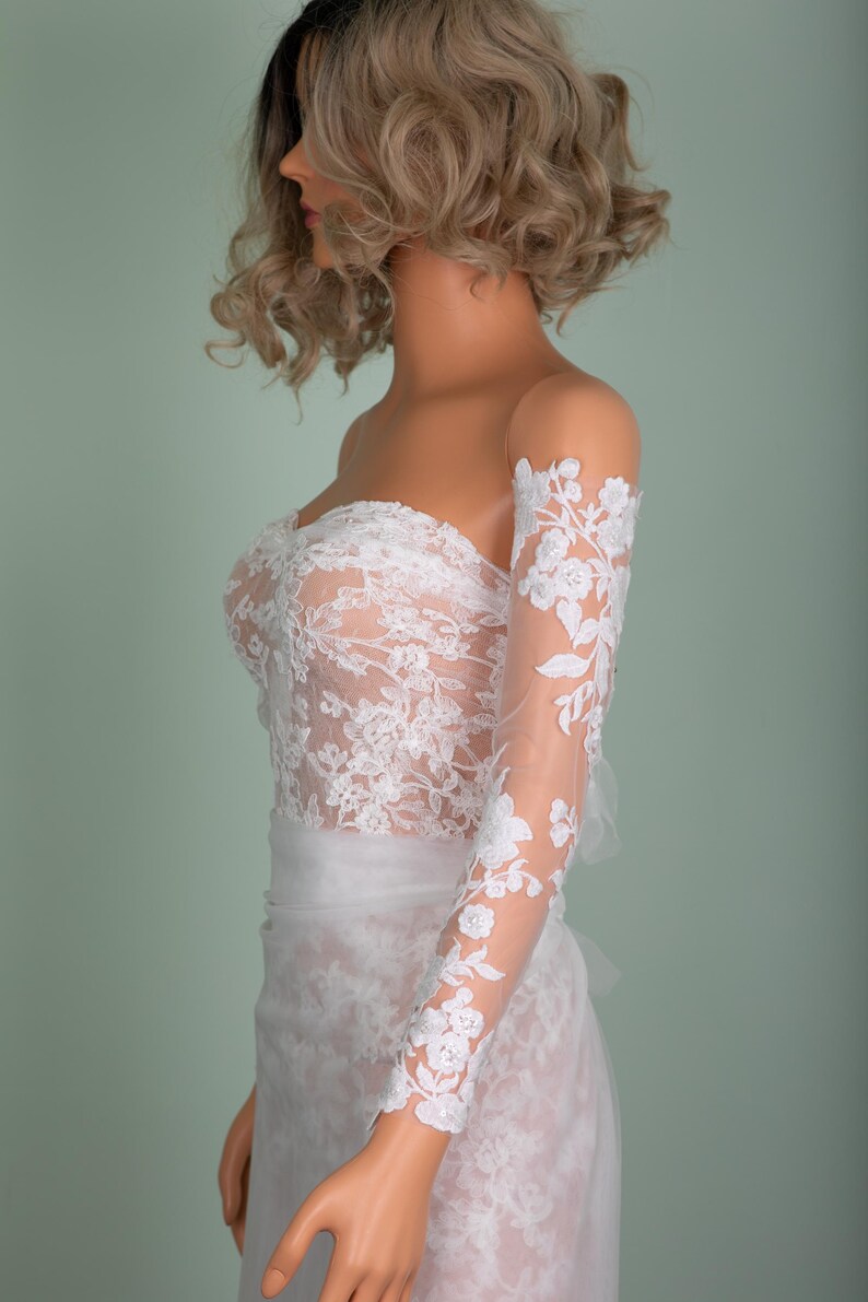 Peut inclure: Robe de mari&eacute;e blanche sans bretelles avec des d&eacute;tails de dentelle florale. La robe pr&eacute;sente un corsage ajust&eacute; et une jupe fluide. Manches longues transparentes avec de la dentelle florale assortie.