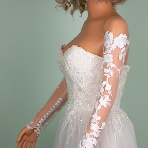 May include: A strapless white wedding dress with intricate lace and beadwork. The dress features sheer, long sleeves adorned with floral lace appliques. The sleeves are buttoned at the wrist, and the dress has a flowing skirt.