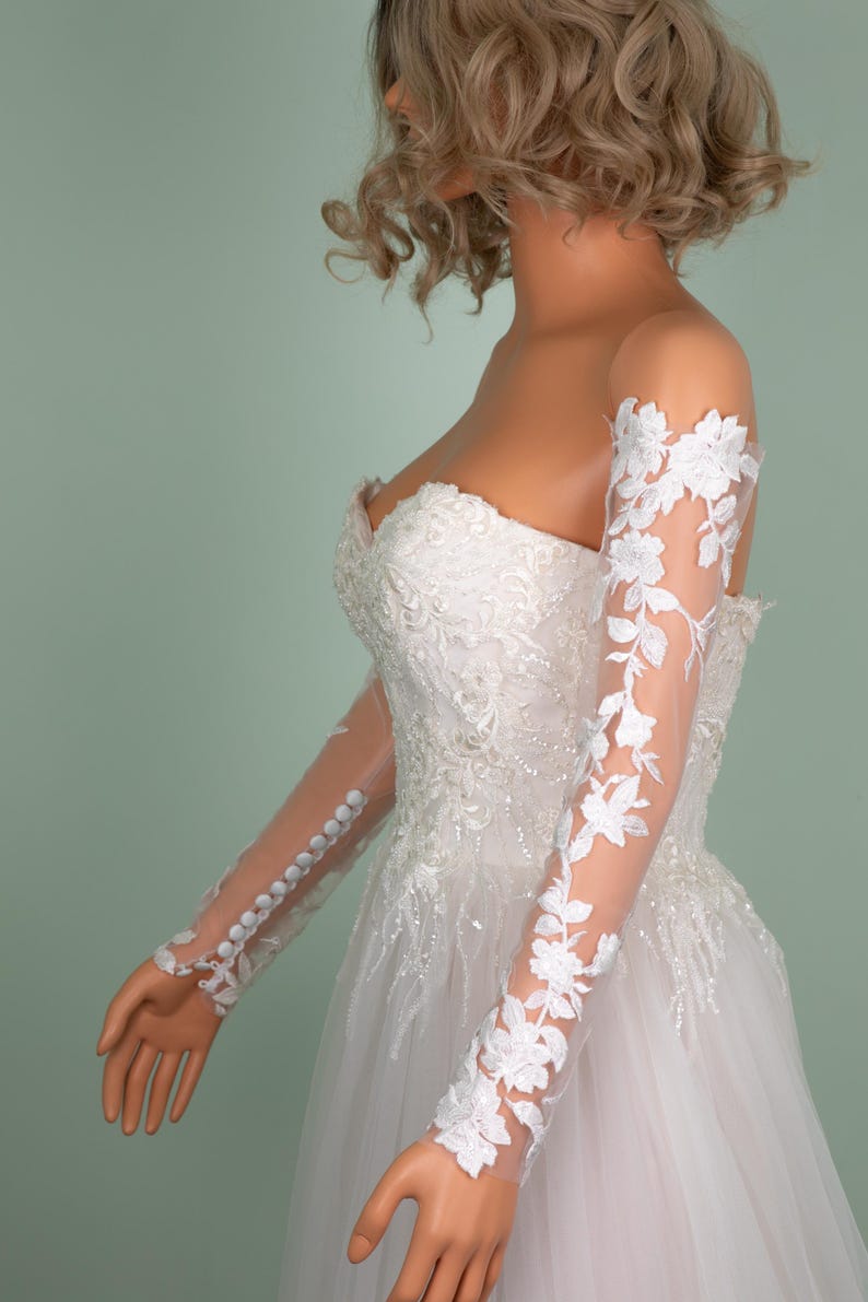 May include: A strapless white wedding dress with intricate lace and beadwork. The dress features sheer, long sleeves adorned with floral lace appliques. The sleeves are buttoned at the wrist, and the dress has a flowing skirt.