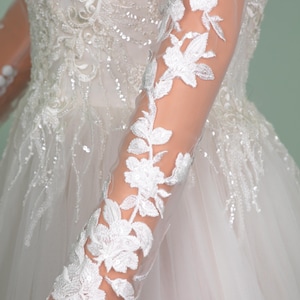 May include: Close-up of a white wedding dress with intricate floral lace detailing. The long sleeves feature delicate white floral embroidery. The bodice is embellished with sequins and beads, adding a touch of sparkle to the bridal gown.