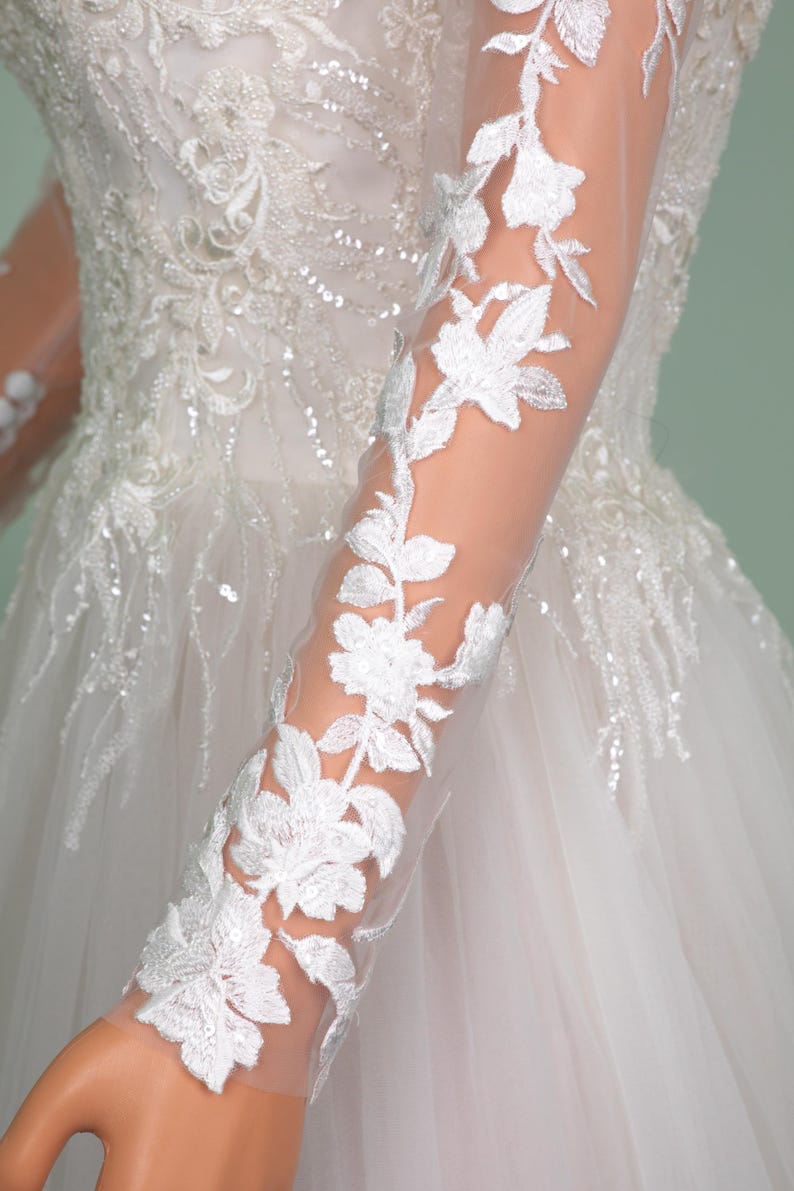 May include: Close-up of a white wedding dress with intricate floral lace detailing. The long sleeves feature delicate white floral embroidery. The bodice is embellished with sequins and beads, adding a touch of sparkle to the bridal gown.