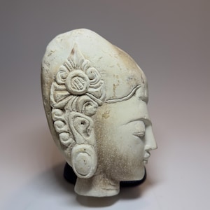 Op de afbeelding: Een zijaanzicht van een uit gebroken wit steen gehouwen sculptuur van een menselijk hoofd. Het figuur heeft gestileerd haar met een ingewikkeld bloemachtig, cirkelvormig ornament aan de zijkant en een glad, langwerpig voorhoofd.