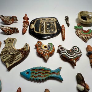 May include: A collection of small, hand-crafted ceramic ornaments featuring various shapes including birds, fish, abstract figures, and a central black rectangular plaque with white circular patterns. The pieces display diverse colors like blue, brown, and green.