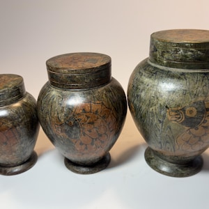 Puede incluir: Tres tarros con tapa graduados hechos de piedra verde oscuro con textura moteada. Cada tarro presenta un diseño tallado de un pez con escamas en tono marrón rojizo, acentuado por contornos oscuros y una base circular.
