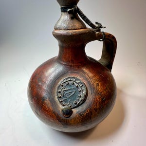 May include: A round, brown, hand-finished ceramic jug with a narrow neck, a sturdy handle, and a removable stopper. The body features a circular, dark-toned medallion with embossed detailing and a small, raised circular accent positioned below it.