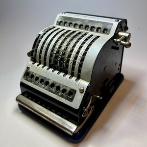 Könnte beinhalten: Ein mechanischer Taschenrechner in Schwarz und Silber mit einer gewölbten Metallfront, die vertikale Zahlenspalten von 1 bis 9 aufweist. Das Gerät verfügt über ein oberes Anzeigefenster mit der Aufschrift "Rosette-BS-9" und eine seitliche Metallkurbel.