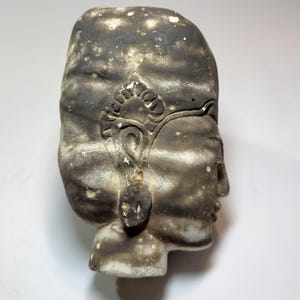 Op de afbeelding: Een grijs en wit keramieken sculptuur van een menselijk hoofd in profiel. Het object heeft een gladde, gevlekte afwerking met donkergrijze schaduwen en een uitgesneden oorornament. De gelaatstrekken zijn subtiel met een duidelijke neus.