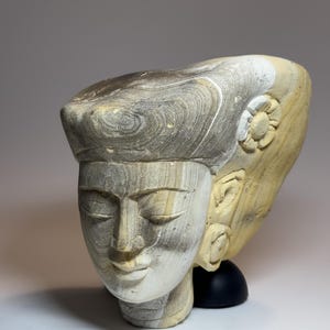 Puede incluir: Una escultura de piedra tallada de una cabeza humana con los ojos cerrados y una expresión serena. La escultura presenta patrones naturales de bandas grises y beige, un peinado texturizado y un pequeño detalle de flor tallada en el lateral.