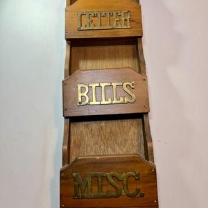 Op de afbeelding: Een verticale houten wandorganizer met drie trapsgewijze opbergvakken. Elk vak heeft een messingkleurige metalen plaat aan de voorzijde met de labels "LETTER", "BILLS" en "MISC" in hoofdletters in reliëf aangebracht.