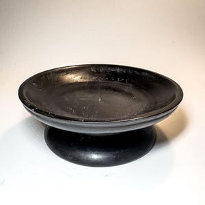 Op de afbeelding: Een matzwarte ronde kom met een brede, ondiepe schaal en een dikke, afgeronde rand. De kom rust op een stevige, voetstukachtige basis die aan de onderkant iets breder uitloopt, wat zorgt voor een stabiele ondersteuning van de verhoogde schaal.