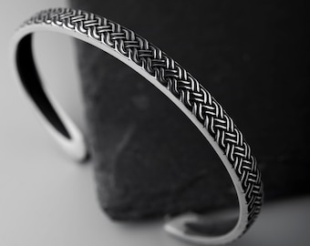 May include: A silver cuff bracelet with a woven design. The bracelet has a textured pattern and is open at the bottom. The jewelry is displayed against a dark background.