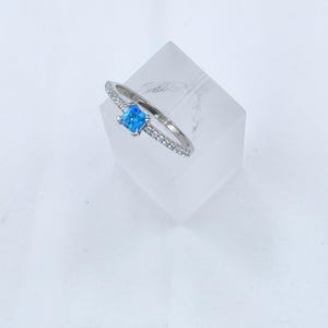 Pode incluir: Um anel de prata com uma pedra preciosa quadrada azul clara no centro. A banda é adornada com pequenas pedras transparentes. O anel é exibido em um suporte geométrico transparente sobre um fundo branco. Este anel é uma joia.