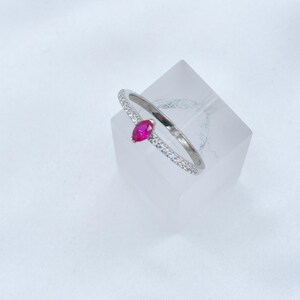 May include: A silver ring with a marquise-cut, bright pink gemstone. The band is encrusted with small, clear gemstones. The ring is displayed on a clear, square block against a white background. Jewelry.