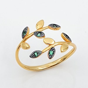 Puede incluir: Anillo dorado con diseño de hojas. El anillo tiene un diseño delicado y abierto con hojas estilizadas. Cada hoja tiene una superficie dorada con un borde oscuro y una pequeña piedra preciosa verde. Es una joya.